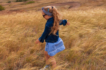 Adorable little girl in denim jacket, black sunglasses, blue plaid dress in yellow grass field. Happy stylish long blonde hair child on countryside landscape. Cute kid walking outdoor rural road trip.の写真素材