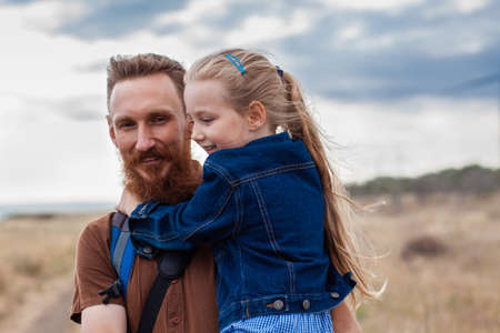 Father with daughter in hands standing on a hill with nature lanscape. Little blonde girl smiling and hugging with dad outdoor. Loving child embrace her daddy. Father's Day family greeting card.の写真素材