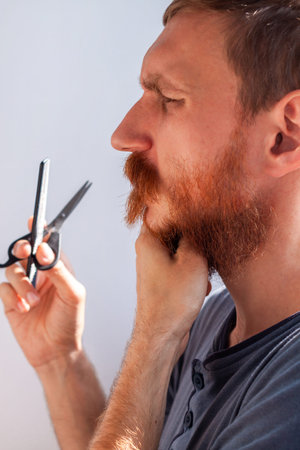 Adult man cutting his own beard and mustache with scissors and comb. Caucasian red bearded male...
