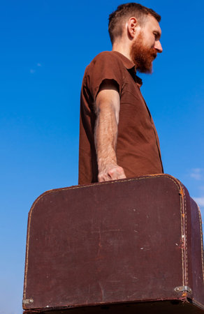 Handsome hipster man with lush beard mustache holds brown vintage suitcase on blue sky background...