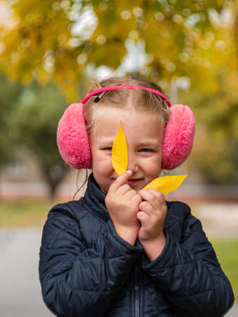 Cheerful little girl in pink fur headphones in autumn park. Smiling young lady walking outdoor in fall. Lifestyle child portrait with blurred natural yellow leaves background. Colorful street photo.の写真素材