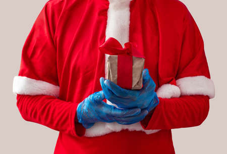 Santa Claus hold offers a gift box with ribbon bow in hands in protective gloves on light background. Christmas shopping, New Year sales mockup copy space. Black Friday COVID-19 safe delivery banner.の写真素材