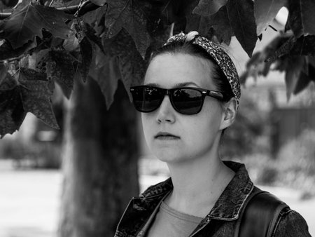 Pretty hipster woman in sunglasses, bandana and denim jacket on nature background on sunny autumn day black and white photo. Positive caucasian young lady smiles side view with copy space. Monochrome.の写真素材