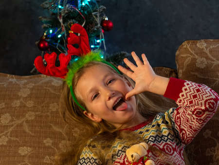Happy little girl in deer horns laughs showing tongue looks into camera at home portrait. Smiling funny toothless child in New Year eve Christmas tree lights. Family holiday celebration spirit conceptの写真素材
