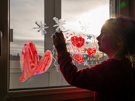 Little girl with paintbrush in hand drawing red heart Love on window glass.の写真素材