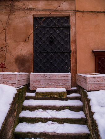 Vintage black metal door with steps in white snow. Antique building exterior detail. Winter street photo retro style. Empty abstract grunge painted texture, weathered brown wall.の写真素材