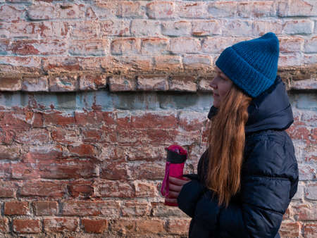 Young woman holds pink reusable bottle of water in hand and drinks on brick wall urban background. Portrait of pretty girl in warm winter clothes on sunny day. Weight loss, active lifestyle body care.の写真素材