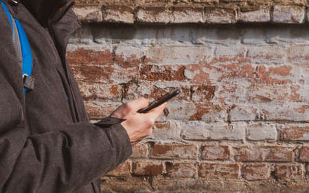 Male hand holding mobile phone and texting. Guy businessman in gray coat on brick wall urban background. Millennial man answers massages using smartphone. Lifestyle distance work communication banner.の写真素材