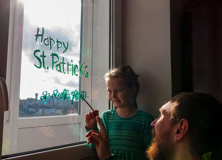 Drawing St. Patrick's Day Father with daughter painting green three-leaved shamrocks indoor, festive home decoration, family leisure. Drawing clover leaves on window glass. Stay home concept New normalの写真素材