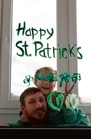 Drawing St. Patrick's Day Father with daughter painting green three-leaved shamrocks indoor, festive home decoration, family leisure. Drawing clover leaves on window glass. Stay home concept New normalの写真素材
