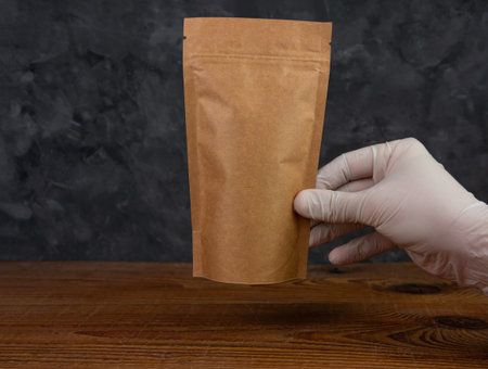 Pouch bag mockup. Hand in protective gloves holds brown paper pack with coffee beans wooden...