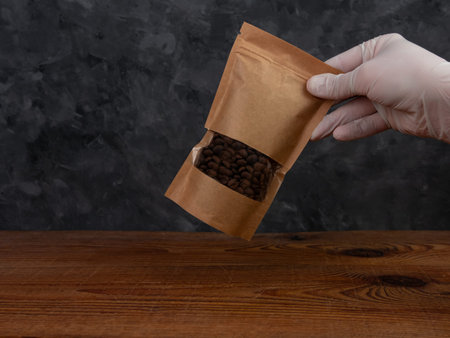 Pouch bag mockup. Hand in protective gloves holds brown kraft paper pack with coffee beans wooden...