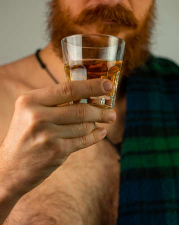 Irish bearded man in traditional green kilt celebrates St. Patrick's Day with drink glass in hand. Scottish guy with mustache drinks whiskey in pub portrait. 17 March parade celebration Irelandの写真素材