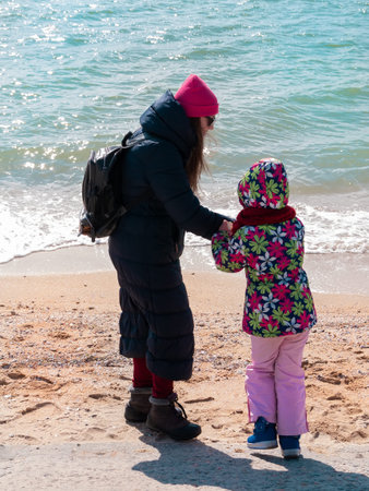 Mother and daughter in warm clothes lifestyle photo on empty beach blue sea and sky. Scenic landscape wallpaper Cold weather spring autumn season. Vivid blue water. Family vacation travel advertising.の写真素材