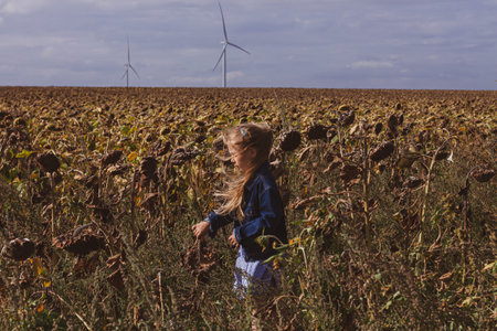 Cute girl in denim jacket showing finger on wind turbines farm on sunflowers field background. Child with long blond hair on countryside evening sunset landscape with yellow flowers. Farming concept.の写真素材