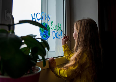 Cute little girl painting planet on window at home. Happy Earth Day April 22 greeting message. Creative family leisure lockdown new reality. Ecology Saving environment conscious consumption concept.の写真素材
