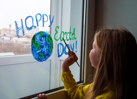 Cute little girl painting planet on window at home. Happy Earth Day April 22 greeting message....