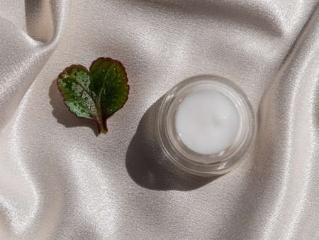Eyelid cream glass jar with green heartshaped leaf on smooth white silk background shadow water splashes sunlight. Natural eco-friendly cosmetic skincare moisturizer skin SPA wellness facial hydrationの写真素材