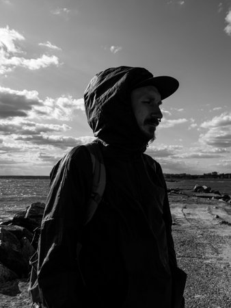 Handsome hipster man with lush beard and mustache on blue cloudy sky background. Red bearded guy in black raincoat with backpack outdoor. Serious brutal male alone. Stylish traveler on empty sea shoreの写真素材