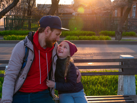 Happy girl with long braided hair and father in spring park sits on a bench on sunset outdoor. Kid...
