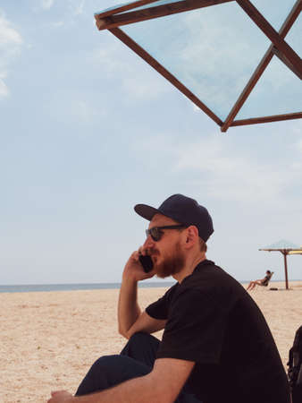 Bearded millennial man in black t-shirt snapback on blue sea landscape with smartphone in hand talking. Young adult guy calling friends on cell phone, real people communication lifestyle distance workの写真素材