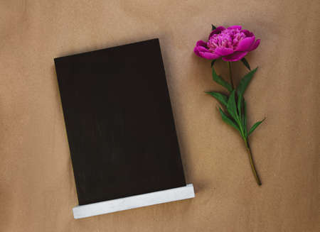 Blank chalkboard mockup with pink peony on brown kraft paper background. Blackboard with beautiful flower wallpaper copy space. Minimalism style. Birthday greeting card floral gift template text sign.の写真素材