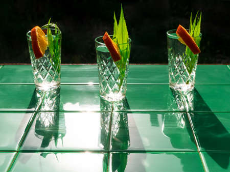 Refreshing drinks in crystal glasses with leaves orange slice on green tile background shadows. Sweet cold natural cocktail hot summer day. Soda water fruits berries mint tonic. Party vacation top viewの写真素材