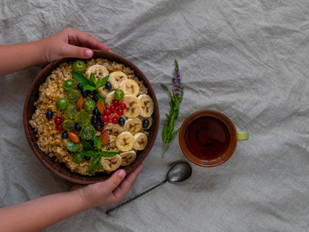Wholegrain Oatmeal porridge bowl with fresh berries fruit nut mint child hands flat lay. Healthy food oat meal morning breakfast top view. Whole grain cereal gooseberry currant banana linen tableclothの写真素材