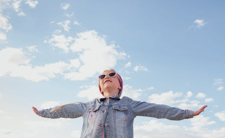 Little blond girl with raised arms blue cloudy sky background. Happy child smiling flying in the...