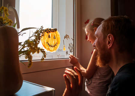 Child with father painting pumpkin on window preparing to Halloween Little girl with dad drawing decorating room paper bats celebration autumn holiday home.Creative family leisure lockdown new realityの写真素材
