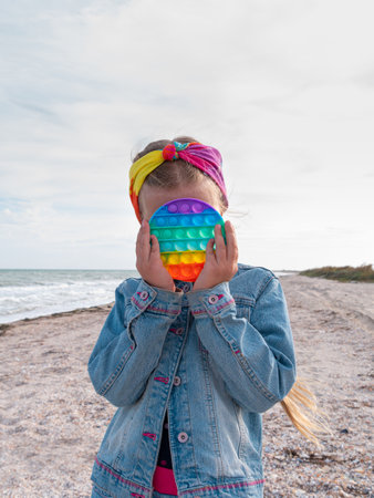 Happy child with rainbow  toy having fun on autumn beach. Blond girl walking on sea background Bright colorful trendy antistress sensory simple dimple fidget push toy in kid hand Lifestyle photoの写真素材