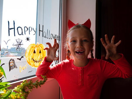 Child in devil horns saying Boo and painting pumpkin on window preparing Halloween. Little kid decorates room interior paper bat celebration autumn holiday home. Creative leisure lockdown new realityの写真素材