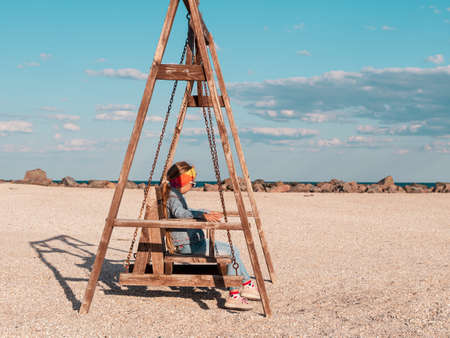 Little girl chilling resting on wooden beach swing. Happy child relaxing enjoying life sun. Lifestyle autumn travel real people outdoor. Having fun fall spring vacation. Freedom happiness adventure.の写真素材