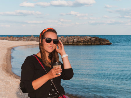 Pretty hipster woman in bandana long hair walking on sea background with coffee cup lifestyle real life travel photo.Millennial girl smiling drinking tea Relaxation Generation Z casual female portraitの写真素材