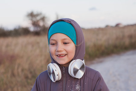 Cute smiling little girl with headphones outdoor on sand beach. Family walking lifestyle. Happy small kid in earphones listening to music and audio books on autumn nature with dry reeds background.の写真素材