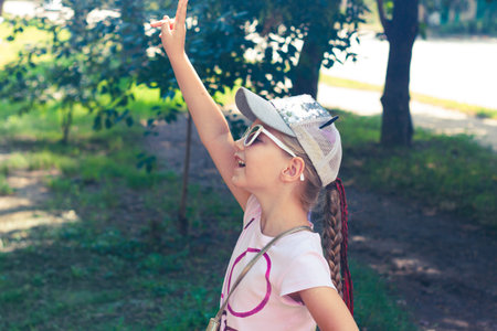 Adorable little girl with long braided hair in baseball cap outdoor on sunny day landscape. Youth culture lifestyle authentic photo. Happy traveler child on green grass in park. School kid walking.の写真素材