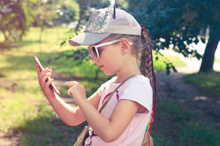 Adorable little girl with long braided hair in baseball cap with smartphone outdoor sunny day. Youth culture lifestyle. Happy traveler child texting in park green grass. Authentic school kid walking.の写真素材