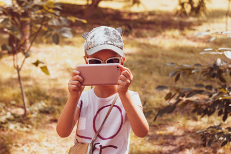 Adorable little girl with long braided hair in baseball cap with smartphone outdoor fall sunny day. Youth culture lifestyle. Happy traveler child texting in autumn park. Authentic school kid walking.の写真素材