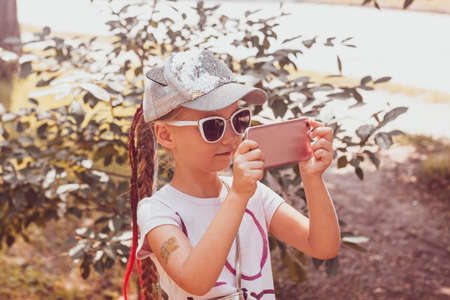 Adorable little girl with long braided hair in baseball cap with smartphone outdoor fall sunny day. Youth culture lifestyle. Happy traveler child texting in autumn park. Authentic school kid walking.の写真素材