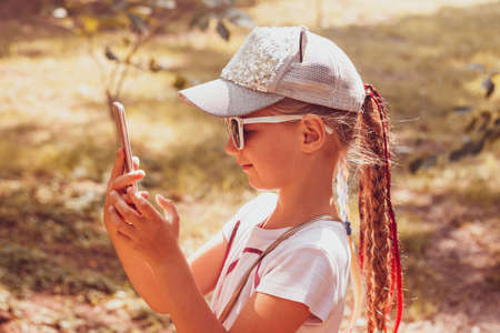 Adorable little girl with long braided hair in baseball cap with smartphone outdoor fall sunny day. Youth culture lifestyle. Happy traveler child texting in autumn park. Authentic school kid walking.の写真素材