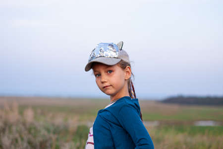 Adorable little girl with long braided hair in baseball cap on a hill outdoor on sunset. Youth culture lifestyle authentic photo. Happy traveler child on countryside landscape. School kid walking.の写真素材