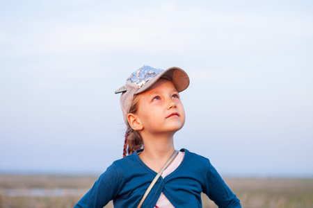Adorable little girl with long braided hair in baseball cap on a hill outdoor on sunset. Youth culture lifestyle authentic photo. Happy traveler child on countryside landscape. School kid walking.の写真素材
