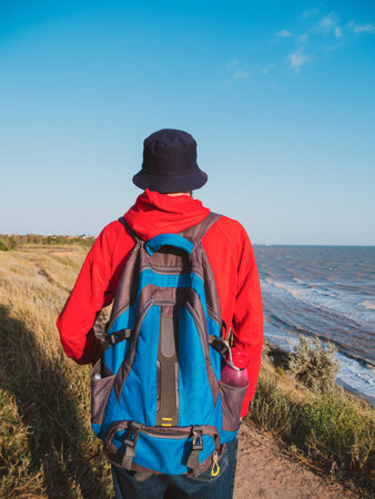 Millennial man in bucket hat red hoody backpack on autumn sea background back view. Authentic male tourist lifestyle photo. Hipster guy outdoor Solo travel adventure concept Active walking backpackingの写真素材