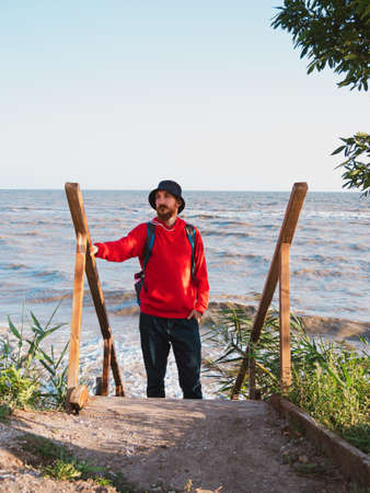 Bearded millennial man in red hoody with backpack autumn sea background dry reeds. Authentic male tourist lifestyle photo. Hipster guy outdoor. Solo travel adventure concept Active walking backpackingの写真素材