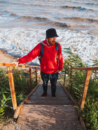 Bearded millennial man in red hoody with backpack autumn sea background dry reeds. Authentic male tourist lifestyle photo. Hipster guy outdoor. Solo travel adventure concept Active walking backpackingの写真素材