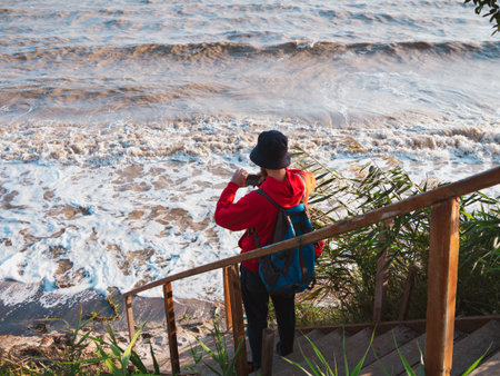 Bearded millennial man in red hoody backpack with smartphone taking photo of autumn sea dry reeds. Authentic male tourist Blogger guy lifestyle Solo travel adventure concept Active walking backpackingの写真素材