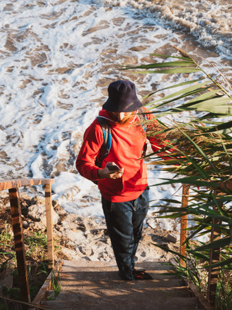 Bearded millennial man in red hoody backpack with smartphone taking photo of autumn sea dry reeds....