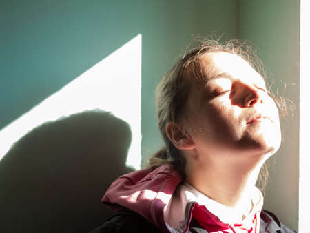 Happy young woman in black raincoat against wall basking in sun autumn day. Smiling girl with closed eyes standing near door sunrays. Lifestyle real people Seasonal depression mental health, Vitamin Dの写真素材