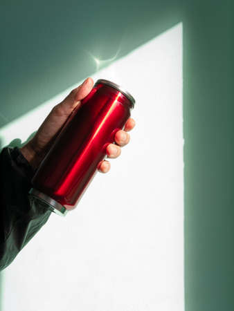 Red thermo can bottle in male hands blue turquoise wall background harsh shadow. Hot tea coffee beverages drinking in cold autumn winter weather. Durable stainless steel reusable eco-friendly thermo.の写真素材