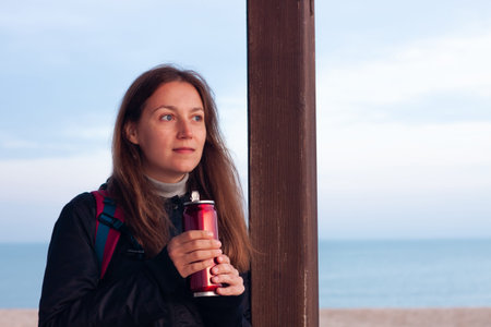 Happy young woman in black raincoat with backpack on empty beach autumn sea. Smiling millennial girl...